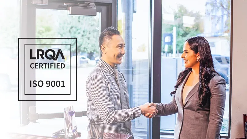 An image of 2 business people shaking hands before a window, and there is the Logo for the LRQA ISO 9001 certification on the left.